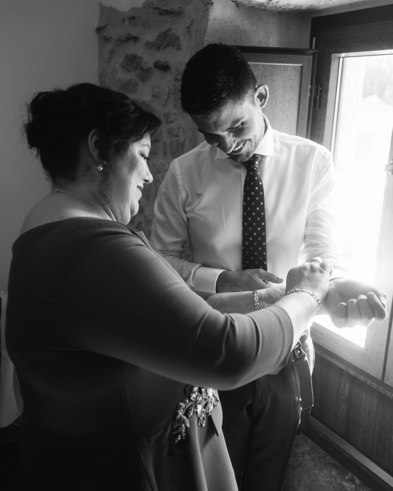 Two people in formal attire stand by a window while helping each other get dressed for a formal occasion.