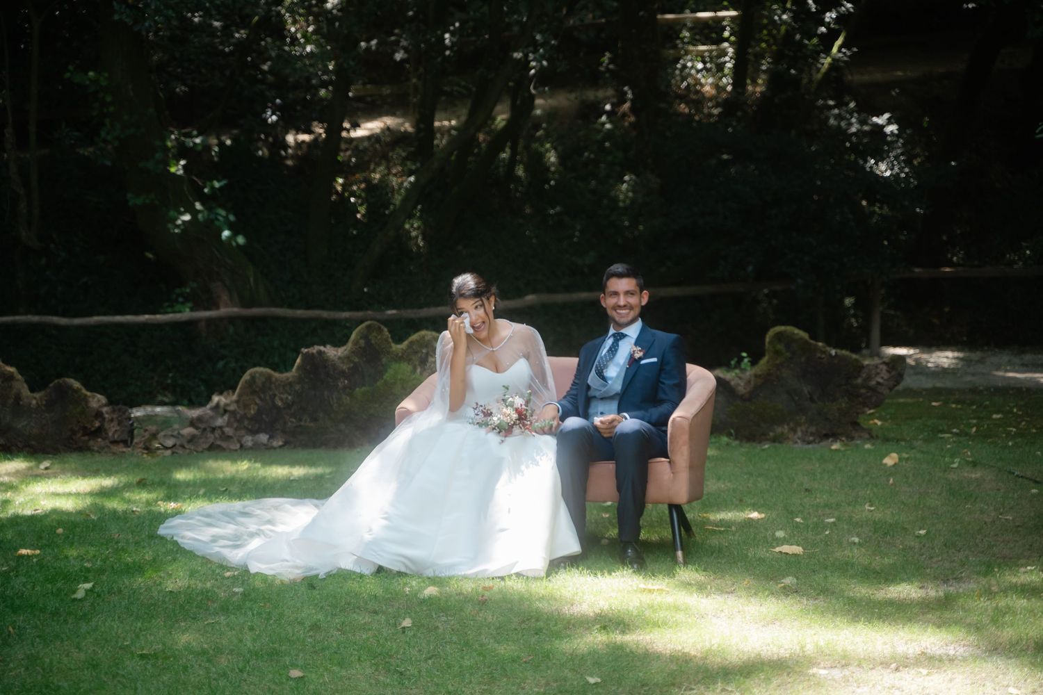 Couple lounges together on a blanket in a sunny garden setting during an outdoor photoshoot.