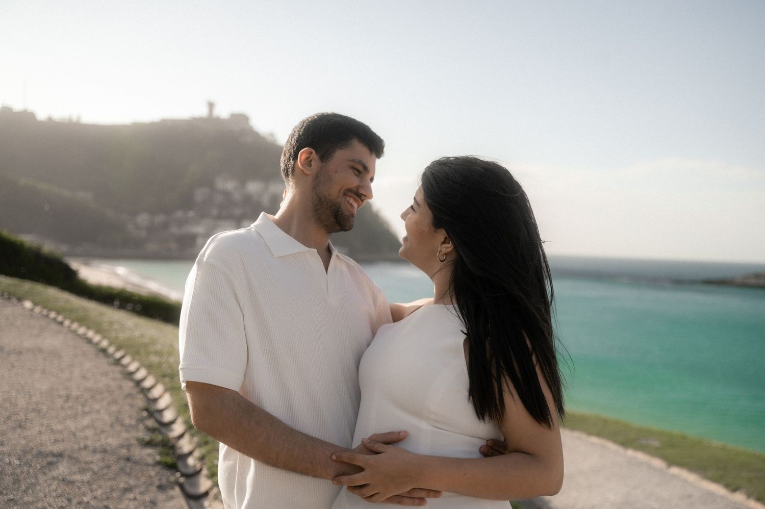 A romantic couple embraces on a scenic coastal cliff overlooking turquoise ocean waters on a sunny day.