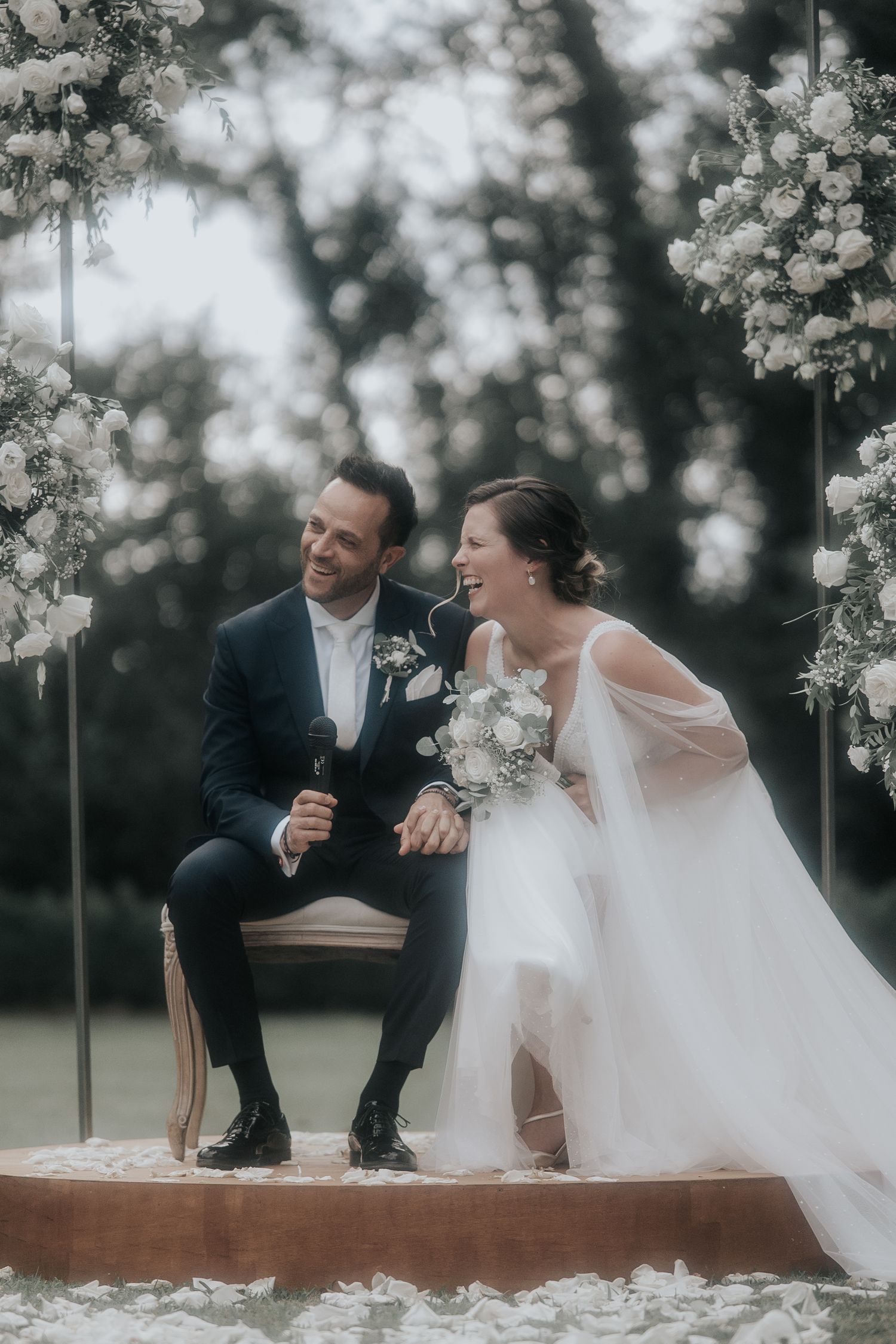 Newlyweds share candid laughter while seated on a rustic wooden platform surrounded by delicate white flowers.