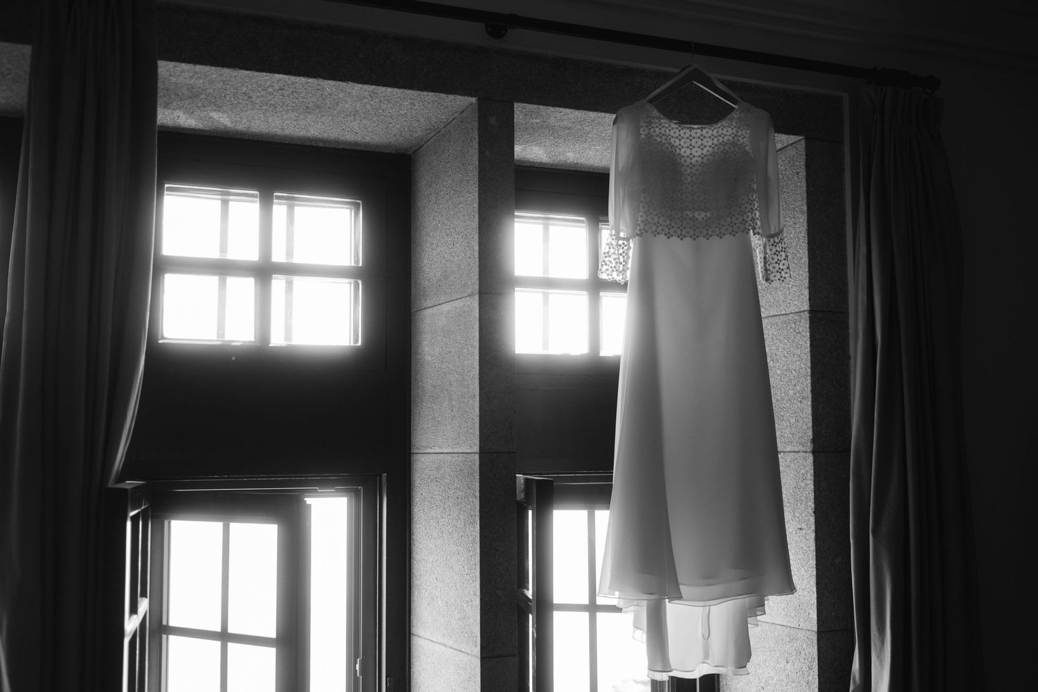 A white wedding dress hangs in silhouette against tall windows in a black and white photograph.