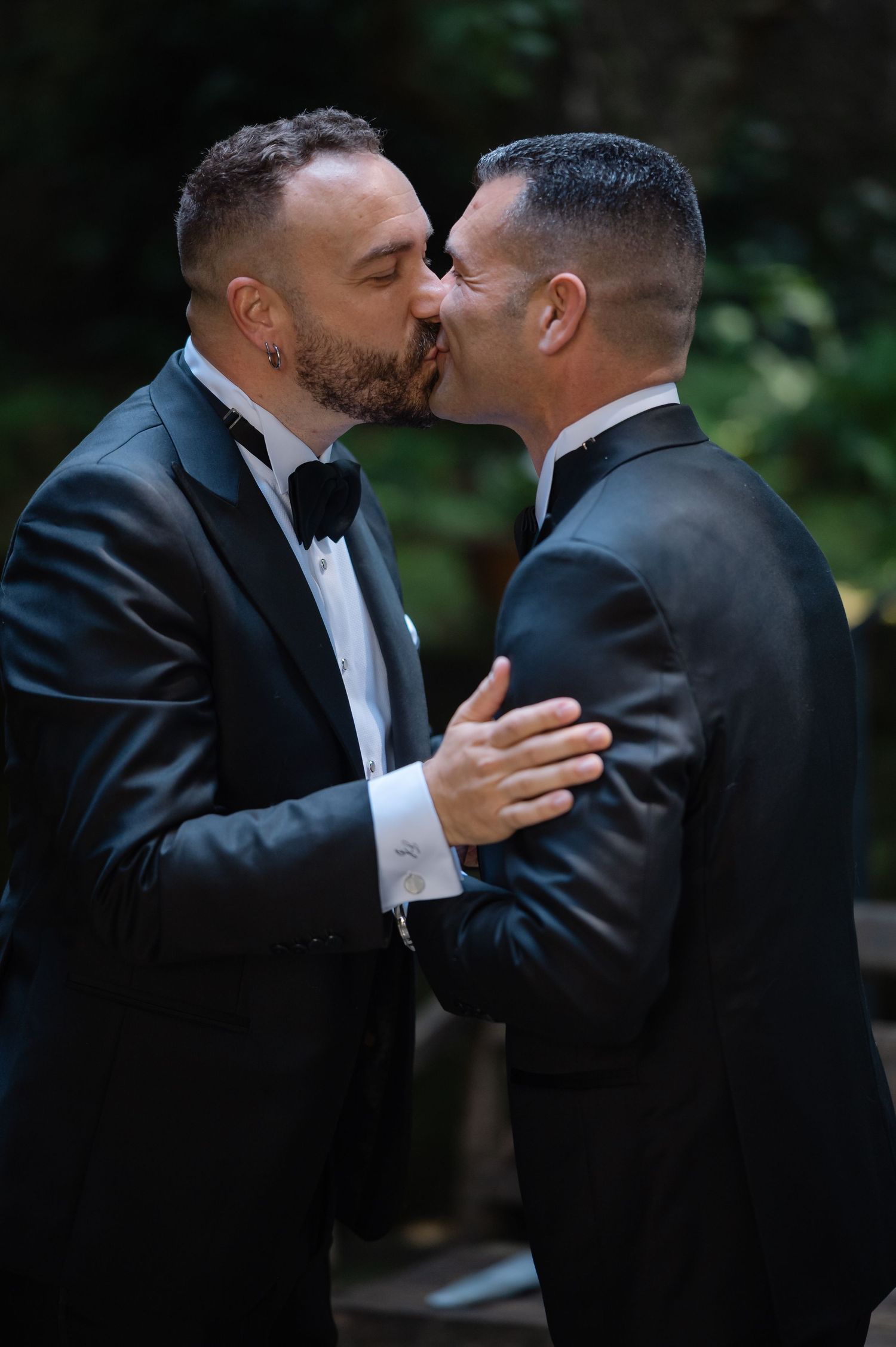 Two people in black tuxedos and bow ties share a romantic kiss during what appears to be a wedding ceremony.
