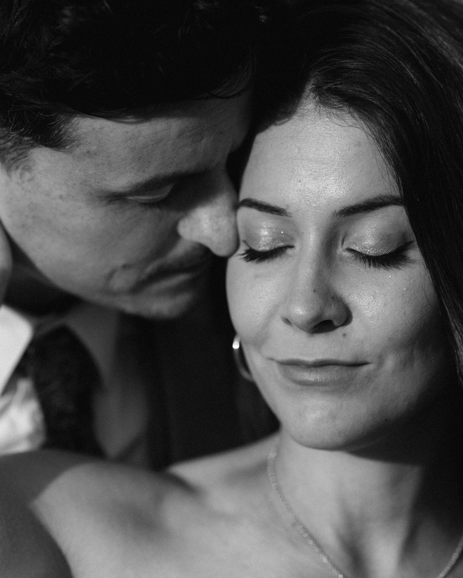 Black and white romantic portrait of a couple, man nuzzling woman's cheek, eyes closed, intimate moment.