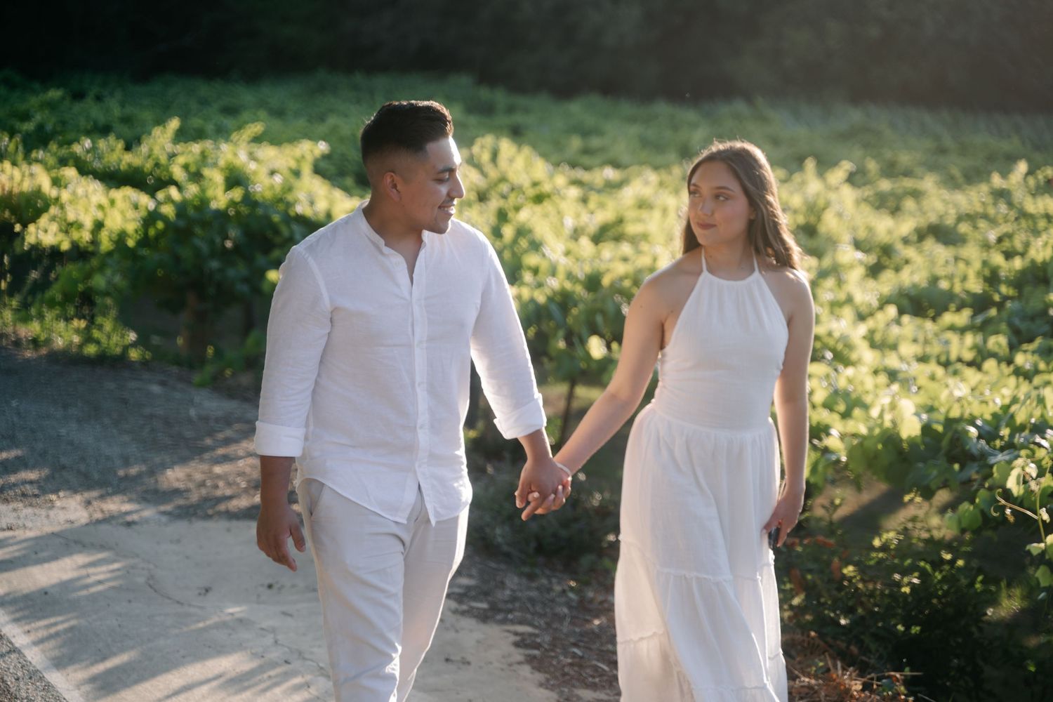 Couple in white outfits holding hands while walking through sunlit garden path.