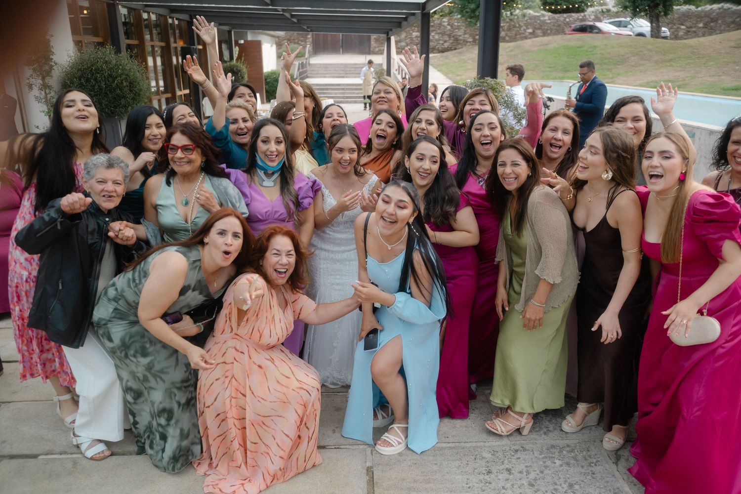 A large group of wedding guests in colorful dresses pose together outdoors for a celebratory photo.