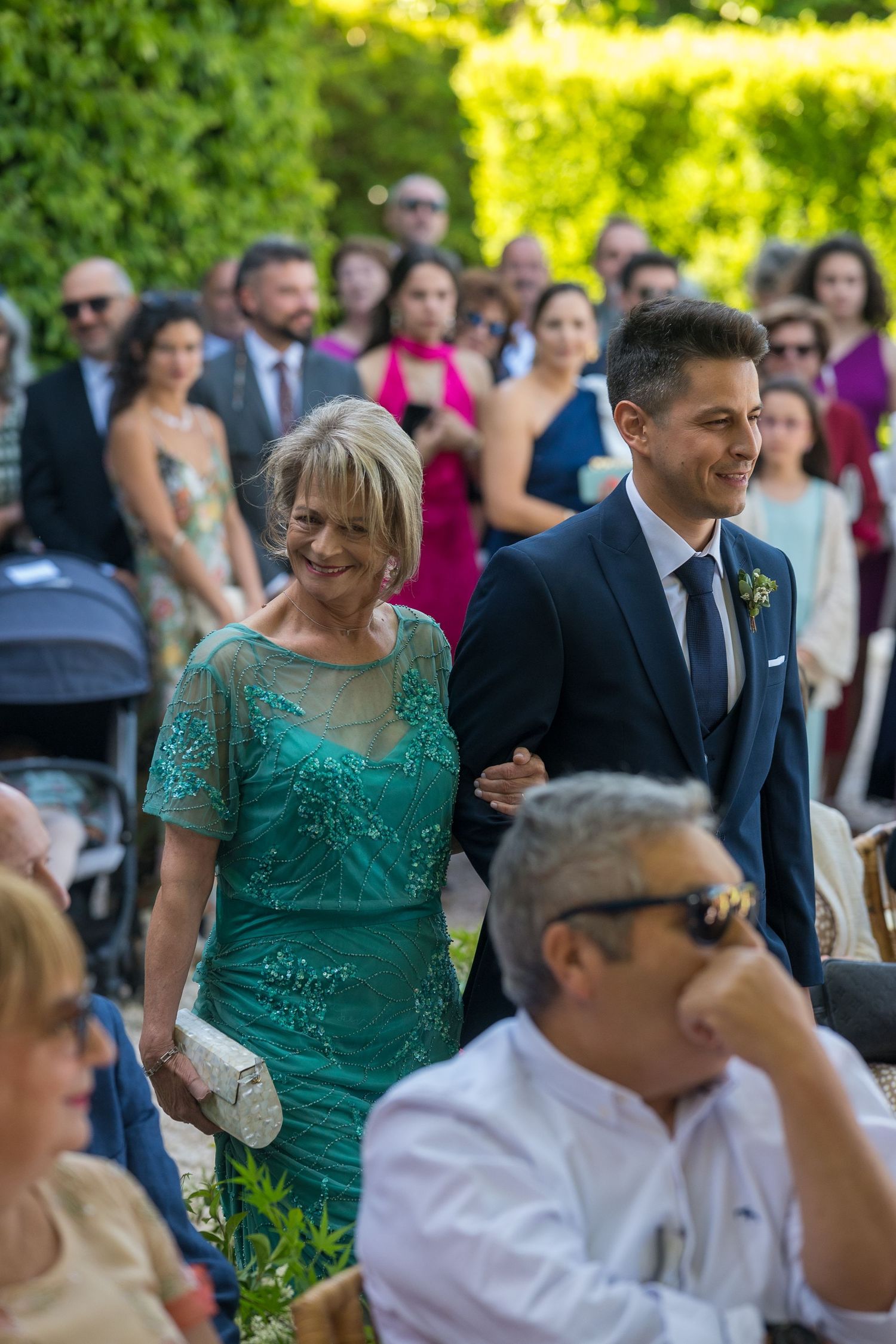 Wedding guests in formal attire mingle during an outdoor reception with lush greenery in the background.
