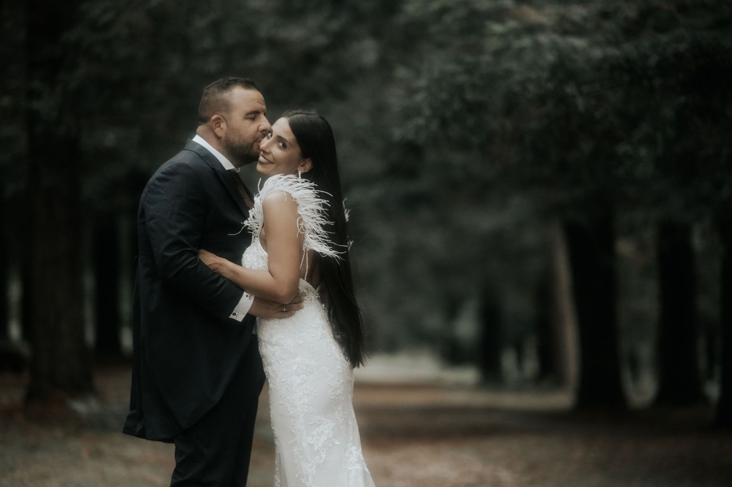 A romantic wedding couple embraces in a moody forest setting with dramatic lighting and dark surroundings.