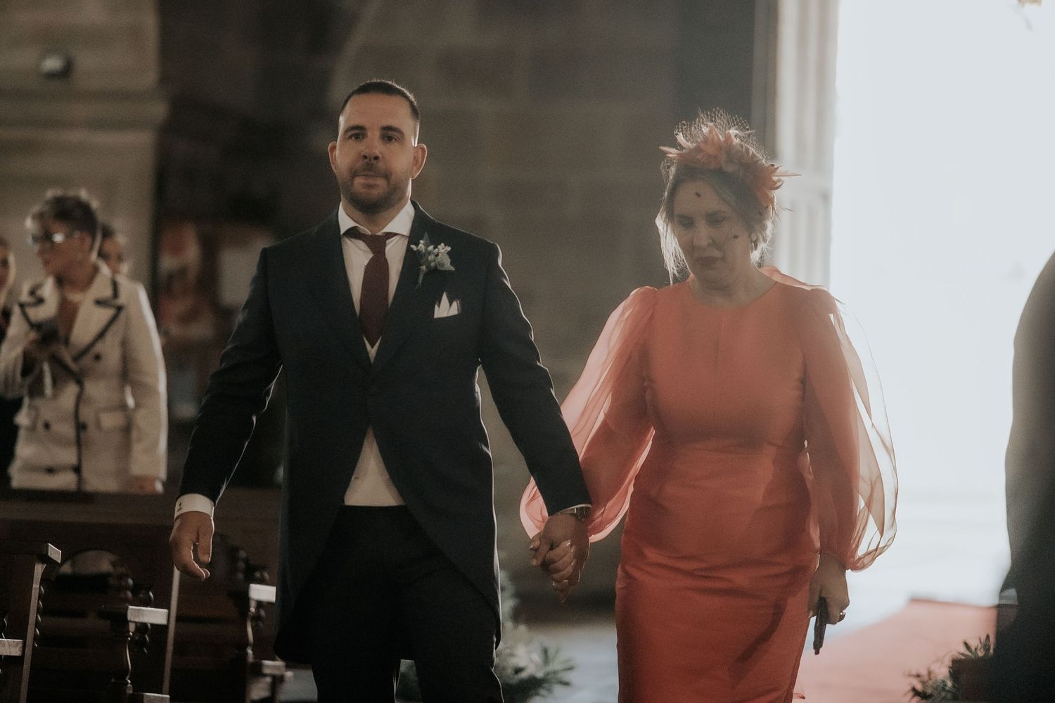 A couple walks hand in hand at a formal event, with the person on the left in a black suit and the right in a coral dress.