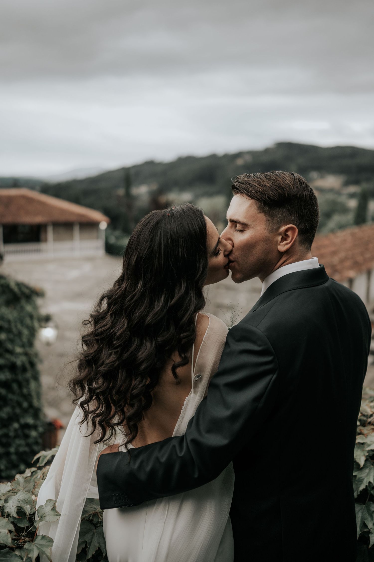 A romantic wedding couple shares an intimate kiss on a cloudy day against a backdrop of rolling hills and terracotta roofs.