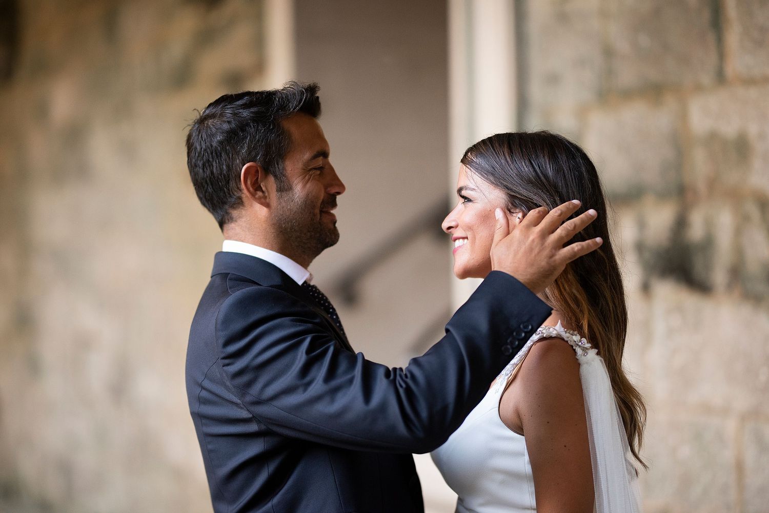 A romantic couple shares an intimate moment in formal attire against a stone wall backdrop.