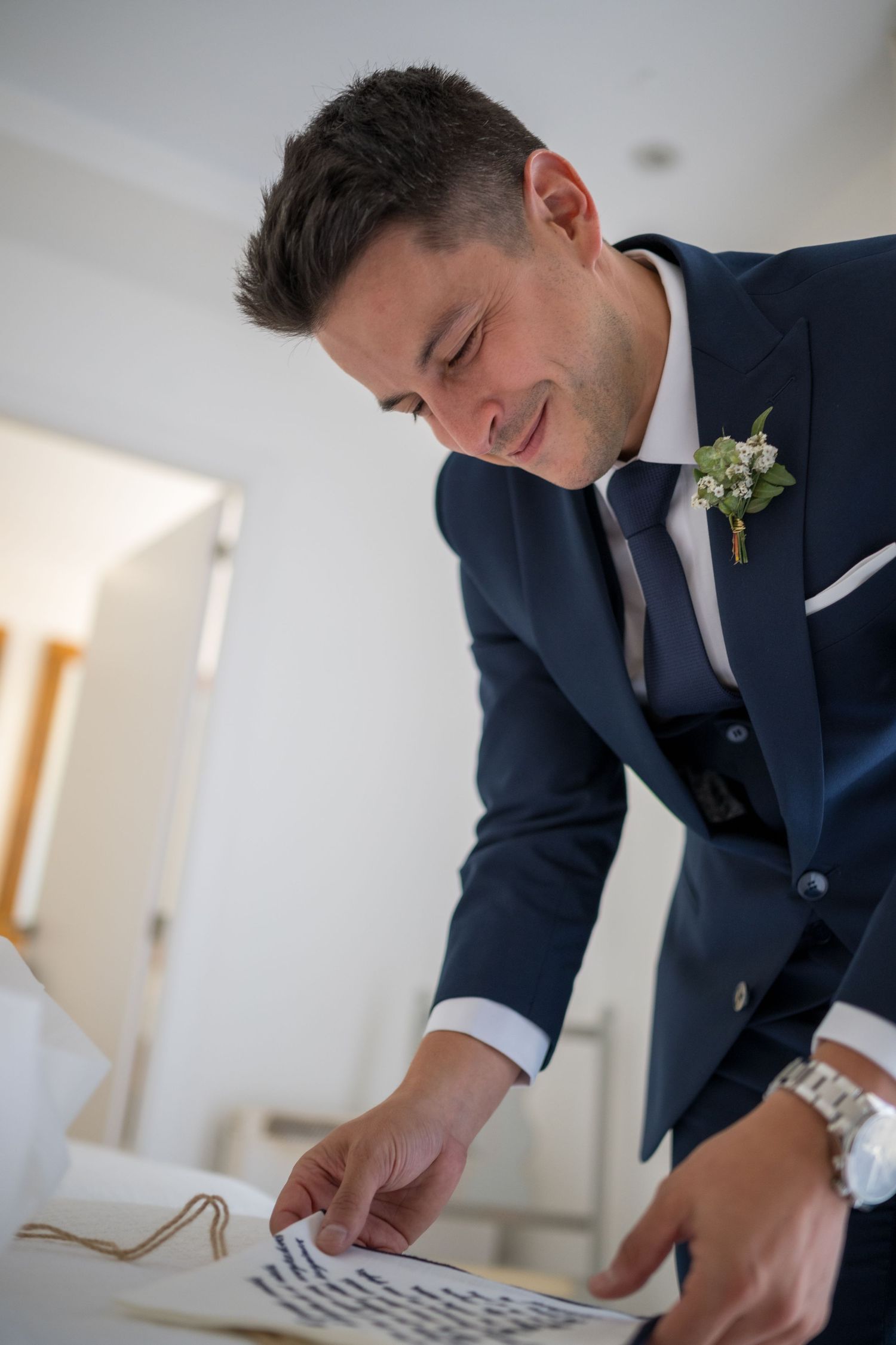 A groom in a navy suit smiles while looking down at papers during wedding preparations.