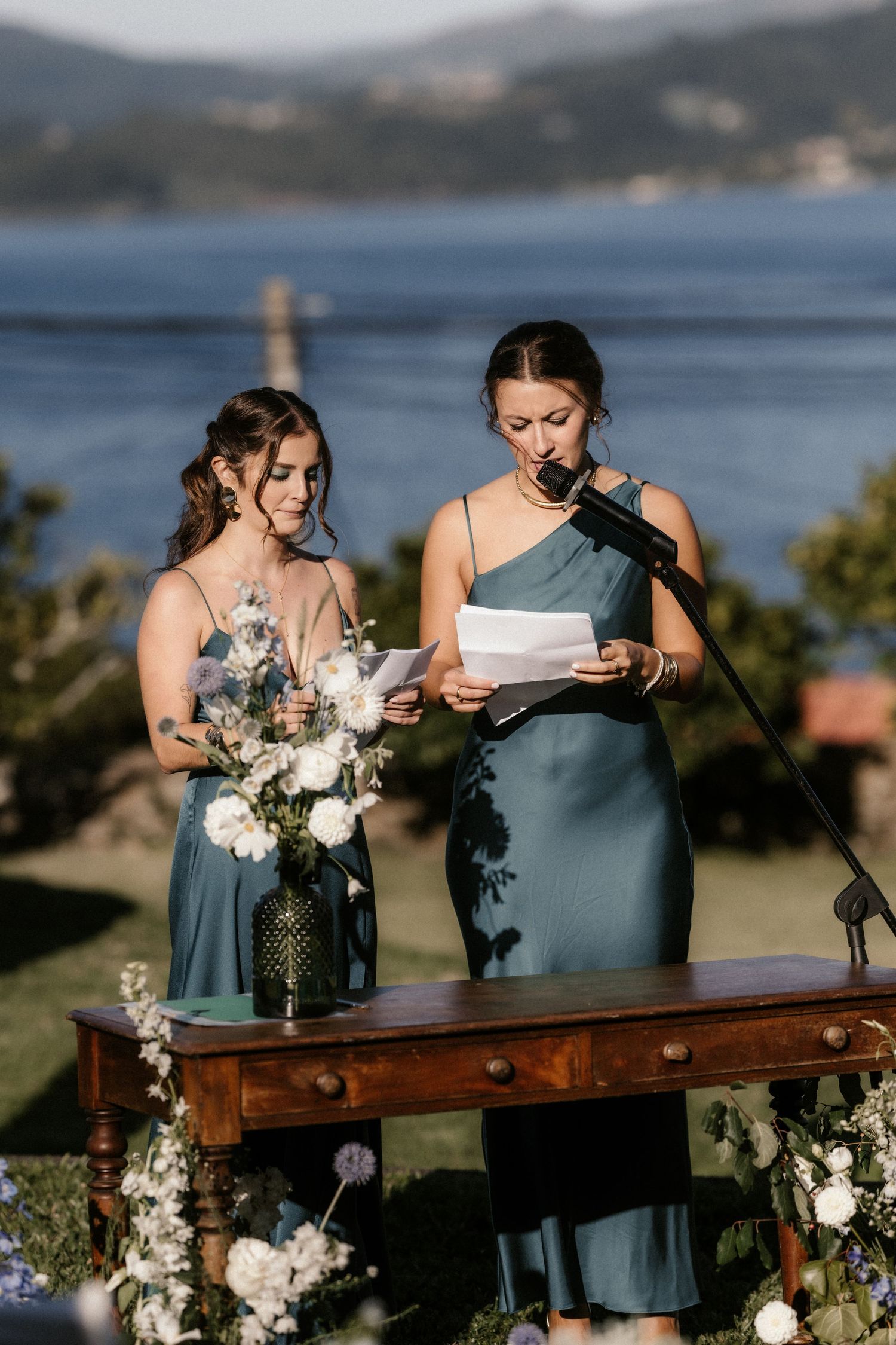 Two people in blue dresses share a tender moment at an outdoor wedding ceremony overlooking a scenic bay.