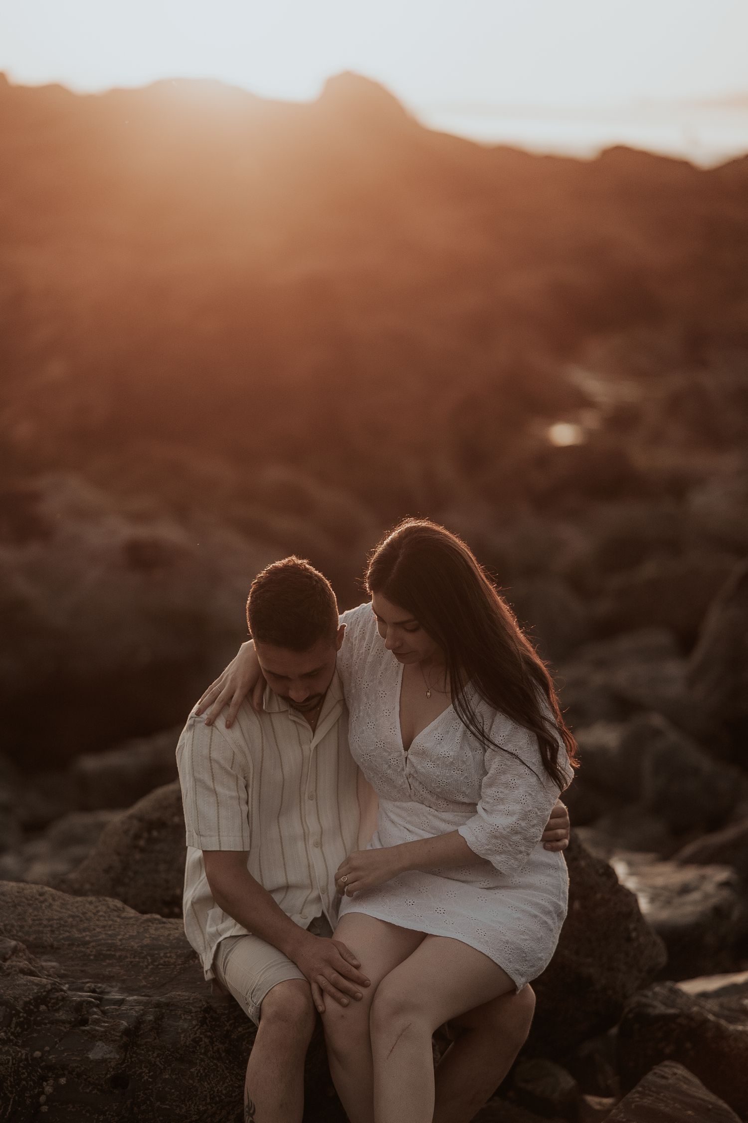 A couple in white outfits sits together on rocky terrain during sunset creating an intimate silhouette.