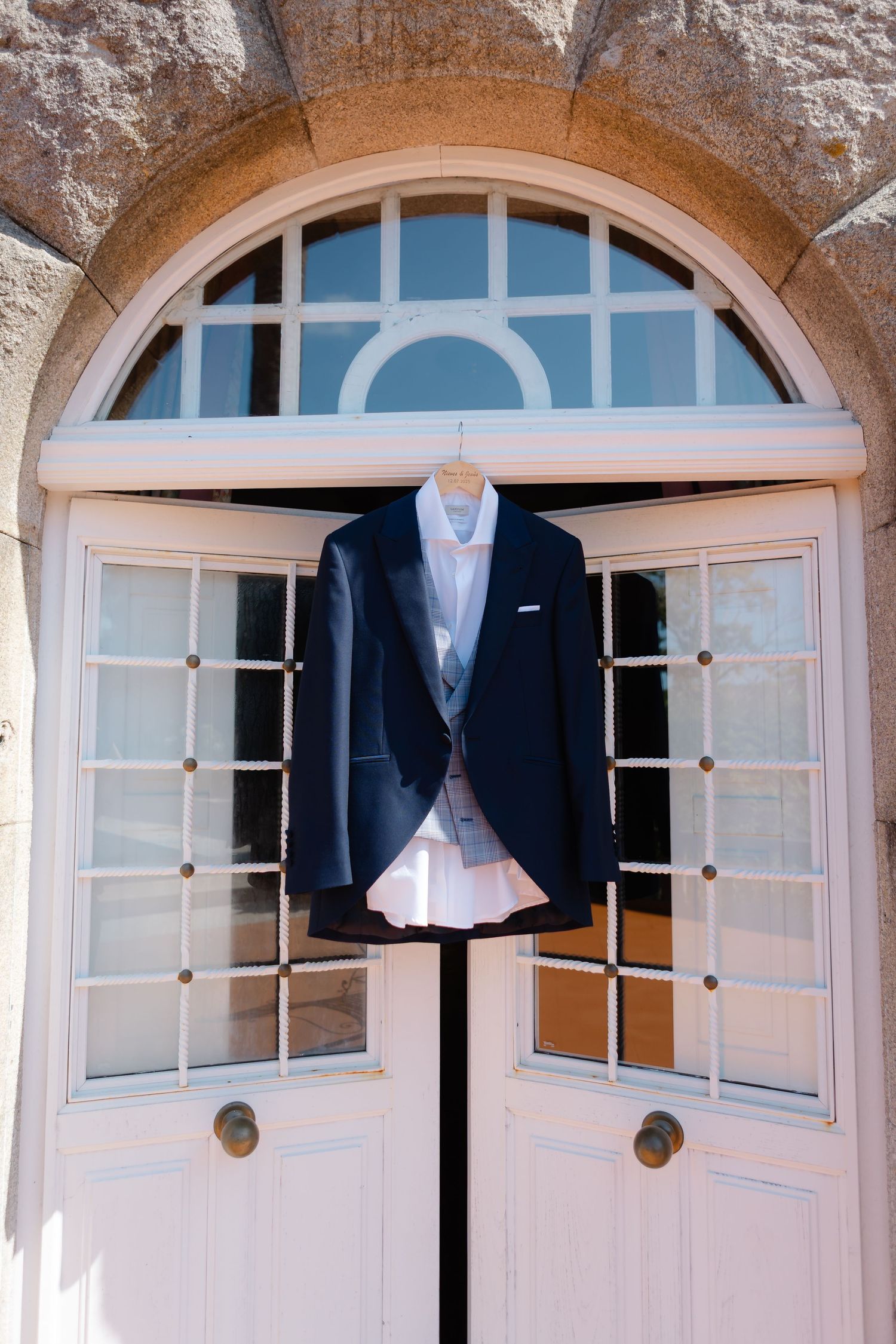 A navy suit jacket and dress shirt displayed on a hanger against an elegant arched doorway with white French doors.