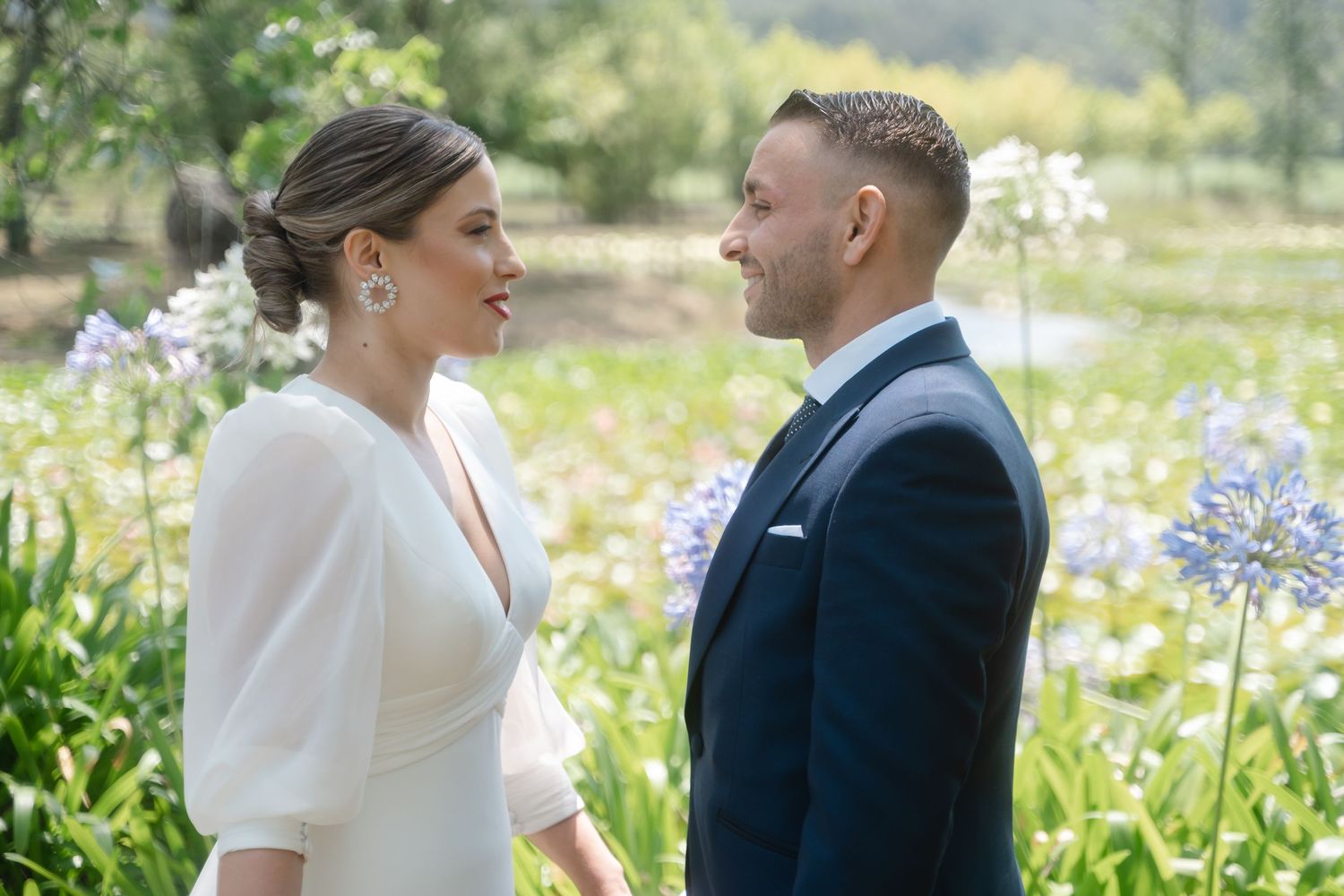 Newlyweds share an intimate moment among flowering plants in a sunlit garden setting.