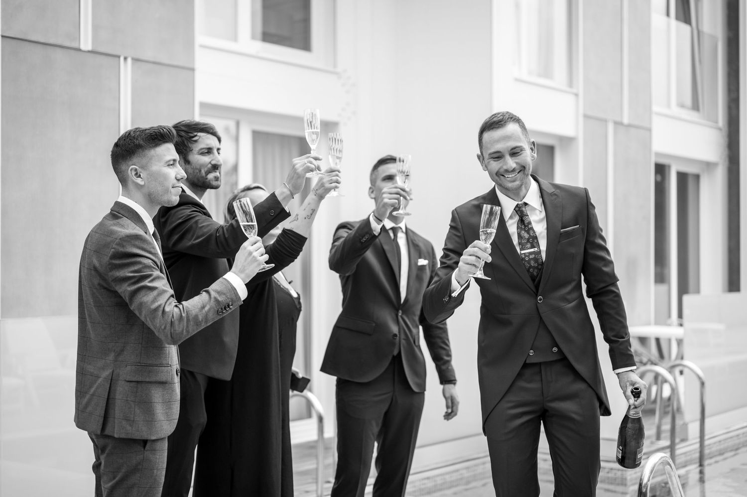 A group in formal black suits and ties stand together in a candid moment outdoors.