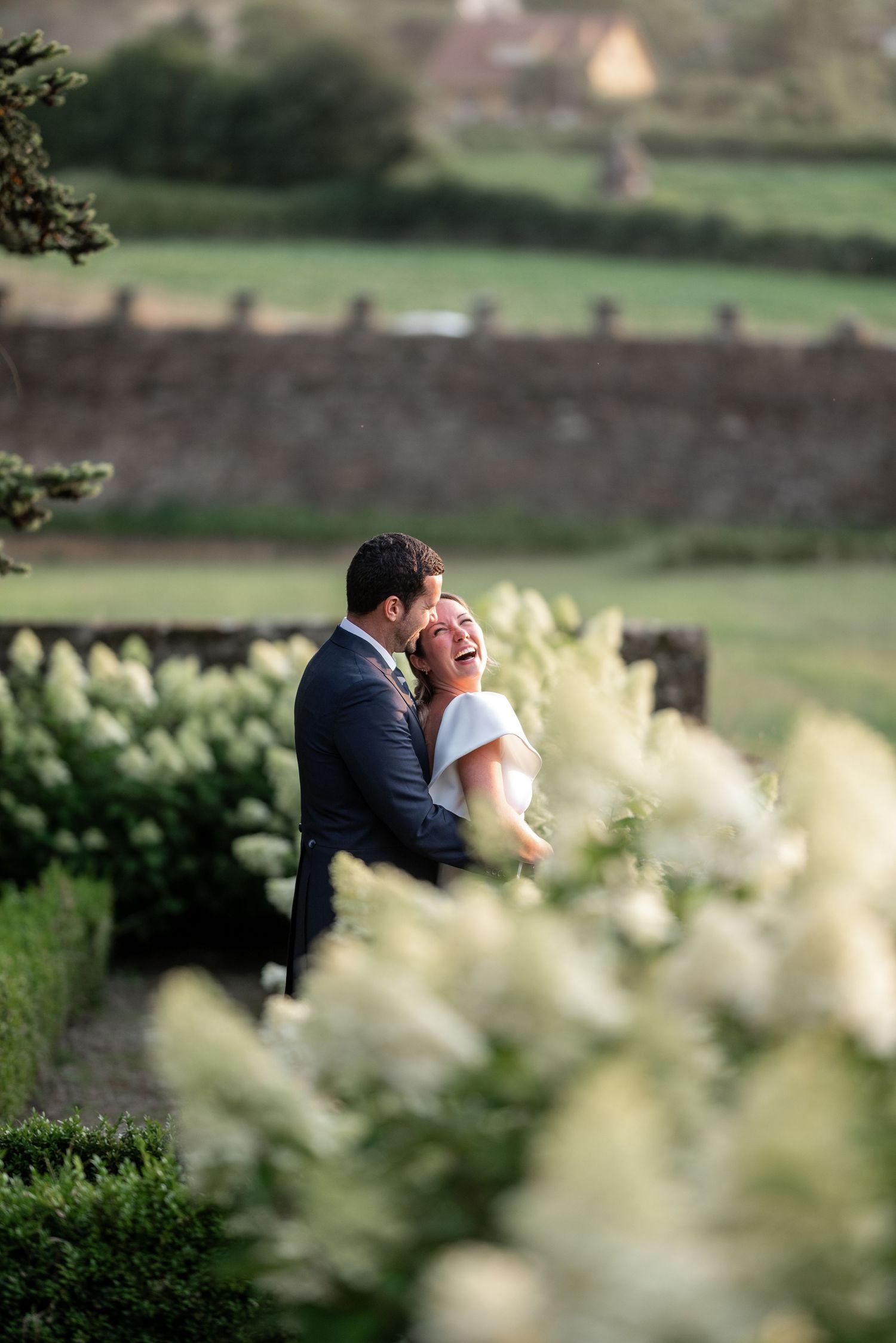 A romantic wedding moment captured through white flowers in a garden with stone walls and green countryside in background.