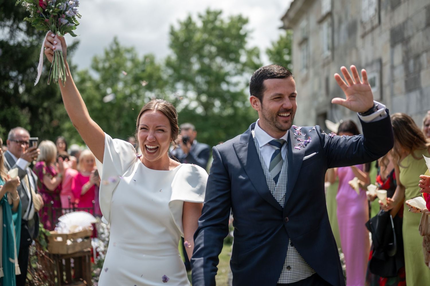 Wedding party celebrates joyfully on a lawn with trees and buildings in the background.
