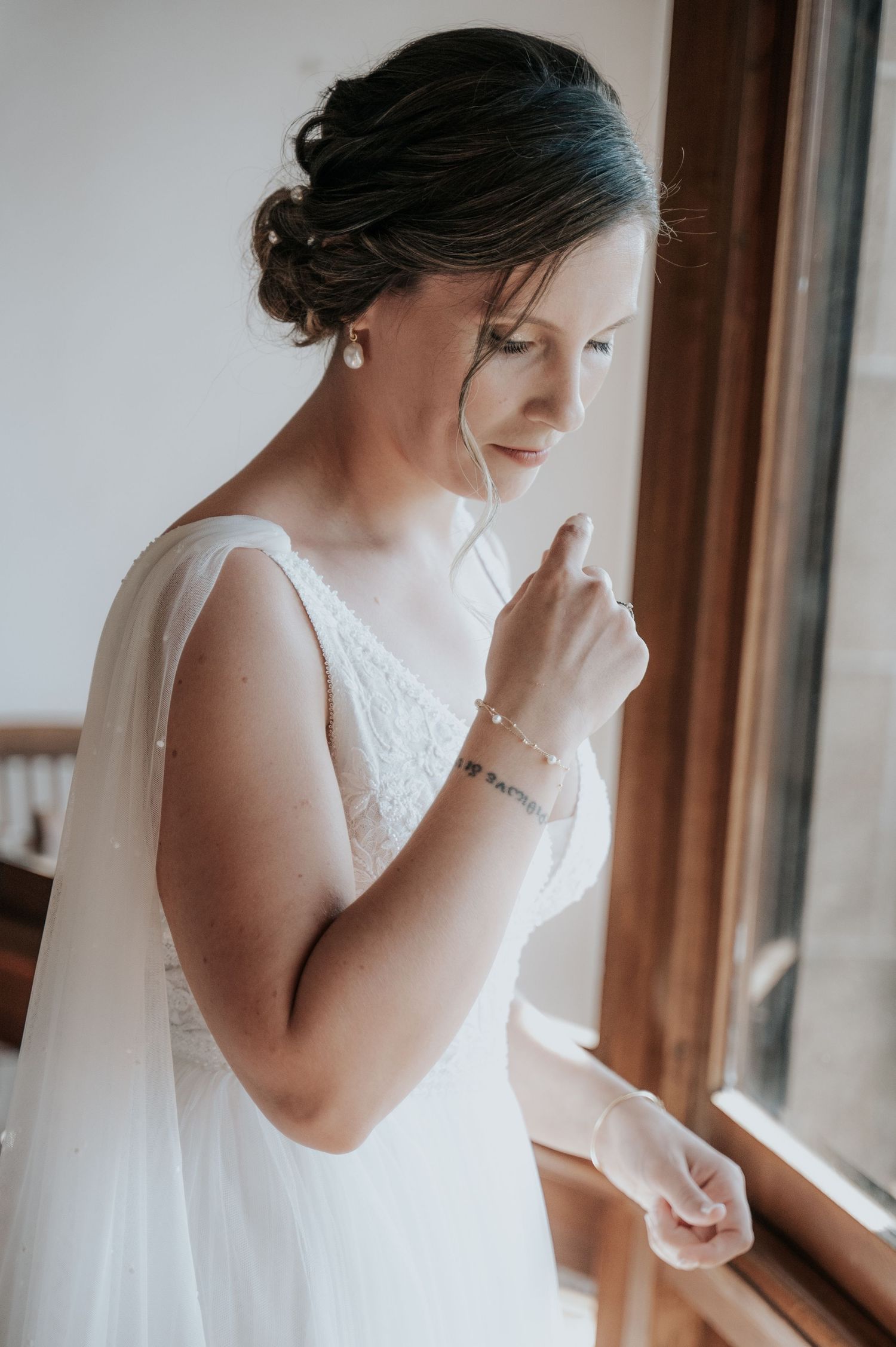 A bride in a white wedding dress adjusts her veil while standing by a window in natural light.