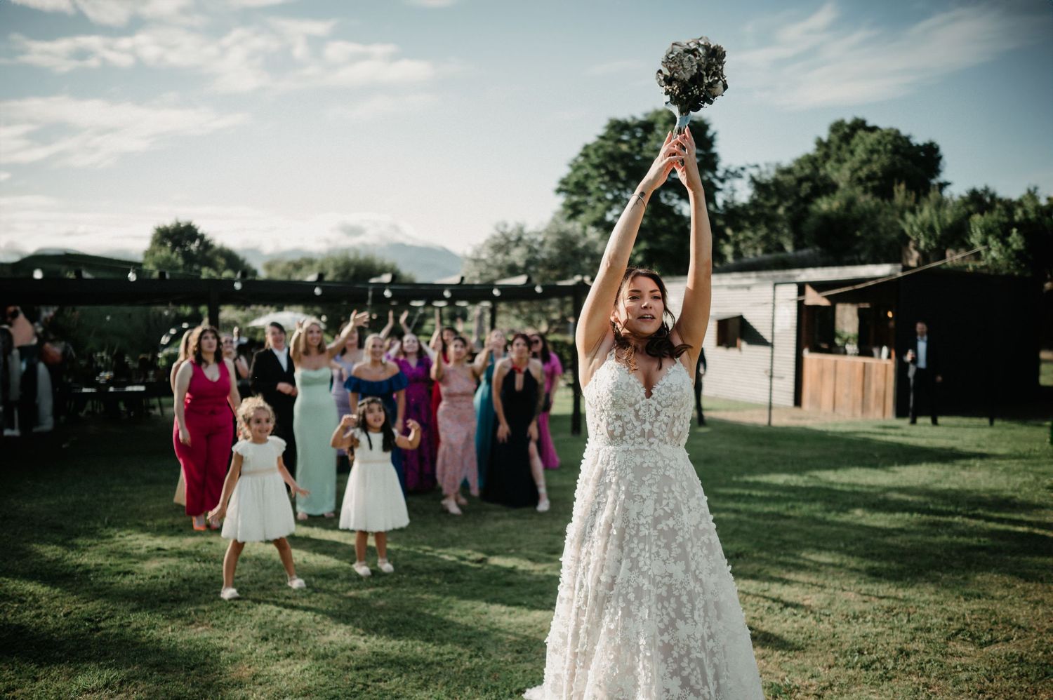 Wedding party gathers outdoors on a grassy lawn during a summer celebration at sunset.