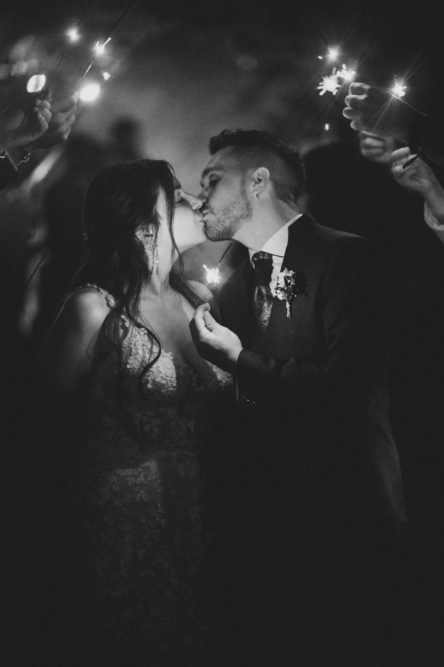 A romantic black and white wedding photo series showing a couple kissing under sparklers at night.