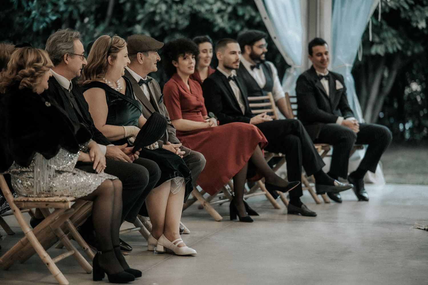 Wedding guests sitting in rows of wooden folding chairs at an outdoor garden venue.