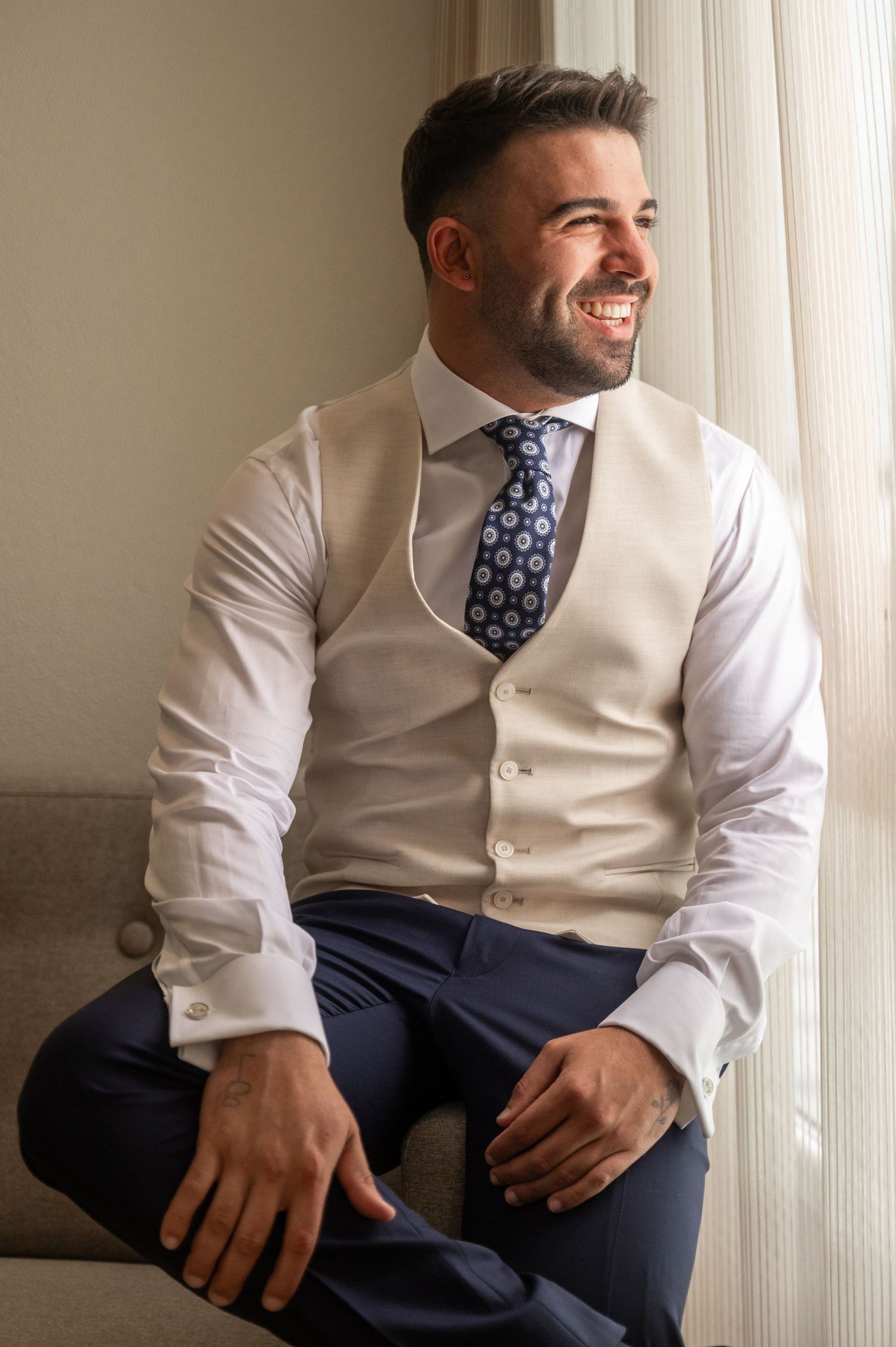 Professional wearing a beige vest and navy tie sits casually by window in business attire.
