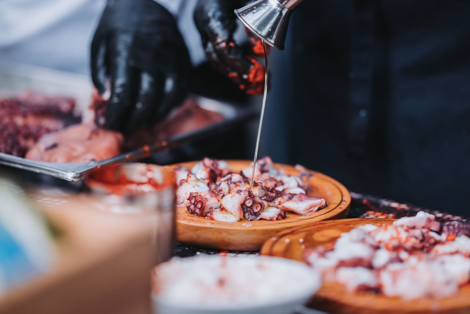 Chef in black gloves drizzles sauce over freshly prepared meat dish on wooden serving board.
