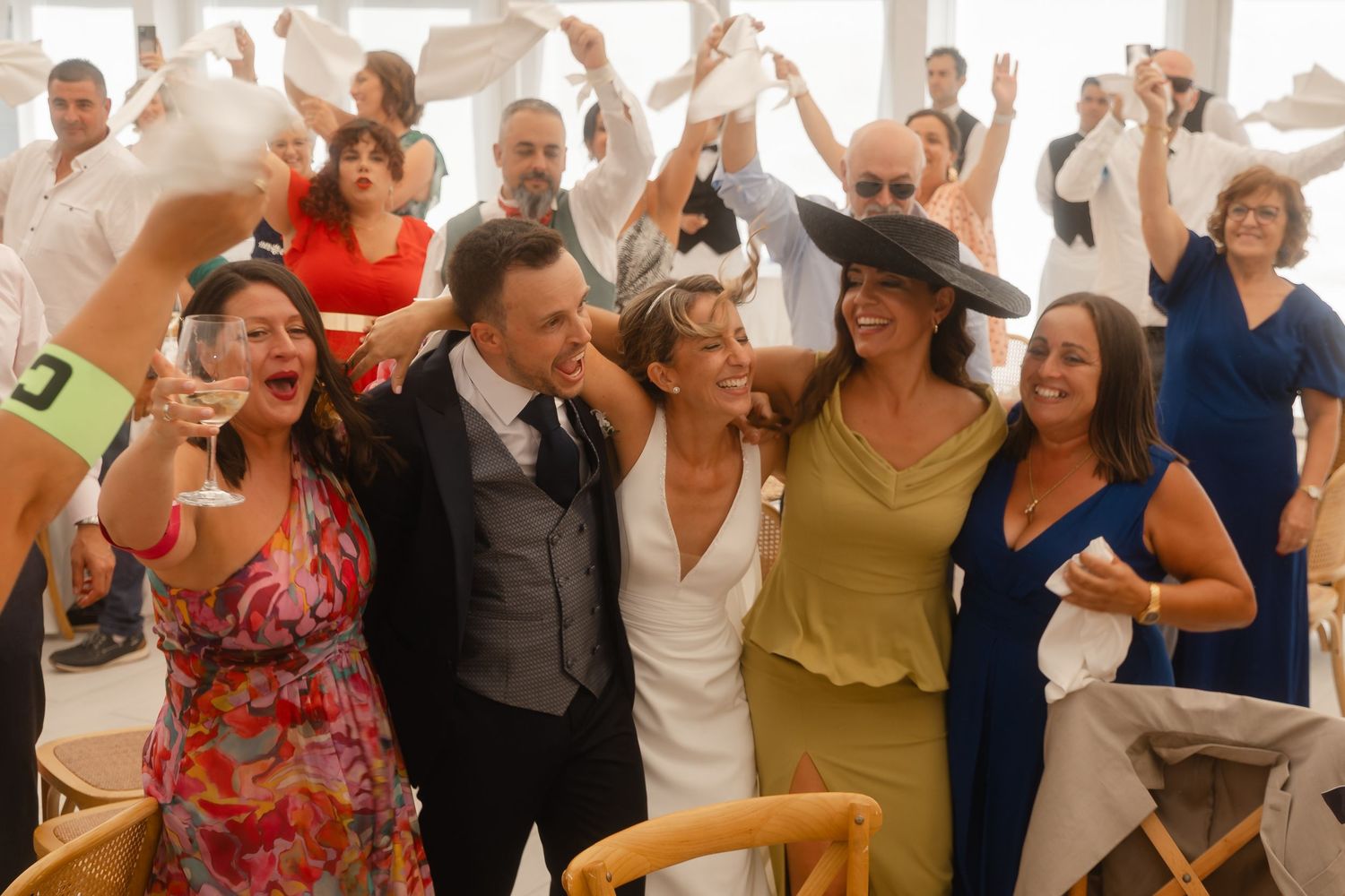 Large group of wedding guests dancing in formation with raised arms during a celebratory reception moment.