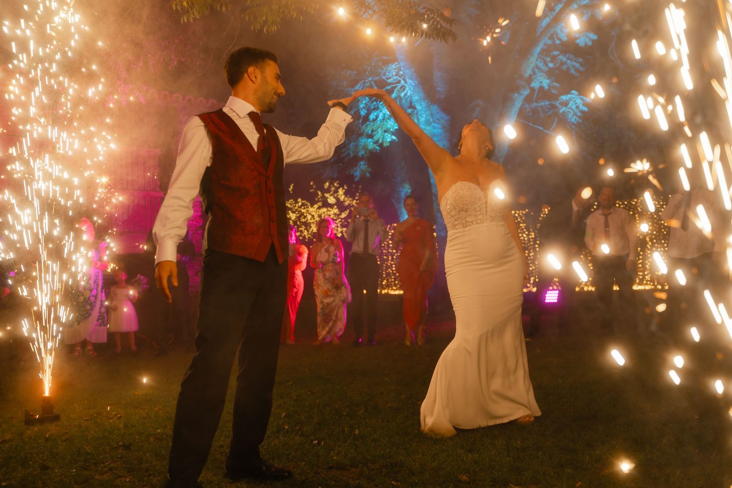 Wedding guests dance under sparkling outdoor lights and fireworks creating a magical nighttime celebration atmosphere.