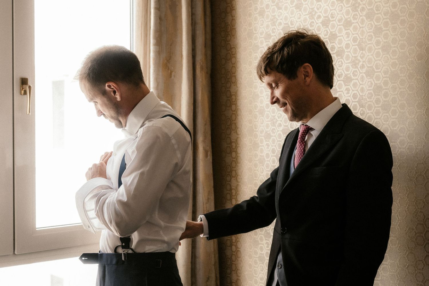 Two people in formal attire stand by a window, with one helping the other adjust their clothing.