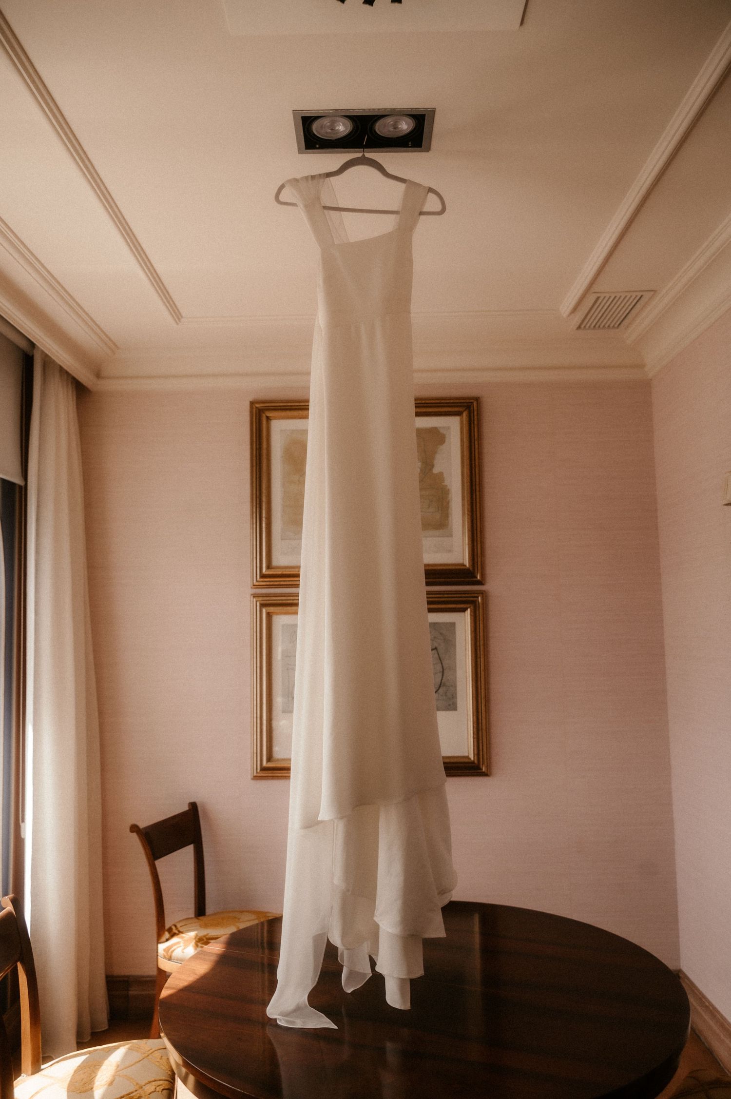 An elegant white wedding dress hangs from a wooden hanger against a light wall with framed artwork and a round table.