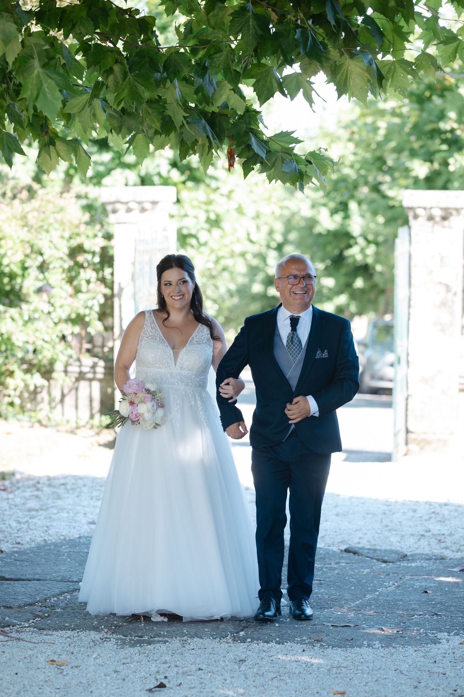 Wedding party walks down a tree-lined path in formal attire during an outdoor celebration.
