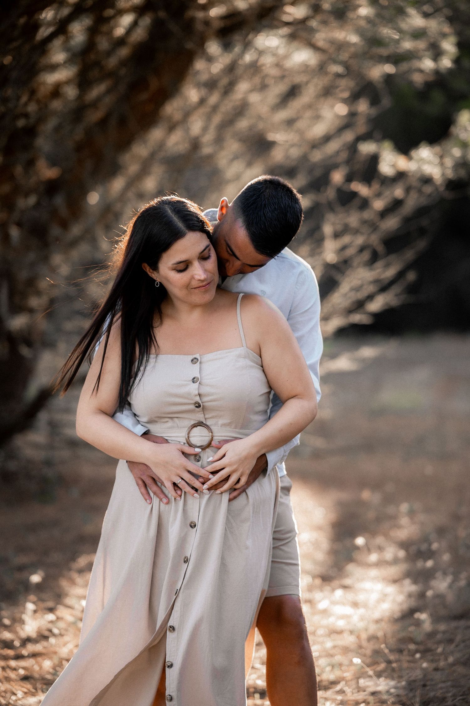 A romantic couple embraces in a sun-dappled forest setting wearing neutral tones during a maternity photoshoot.