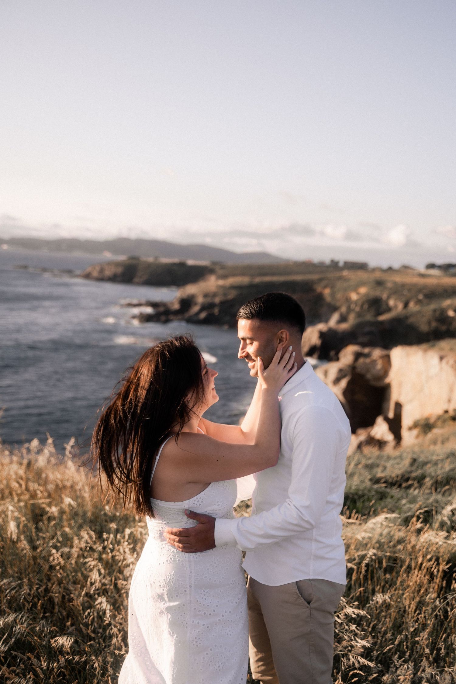 A couple embraces on a scenic coastal cliff with dramatic ocean views and rugged terrain at sunset.