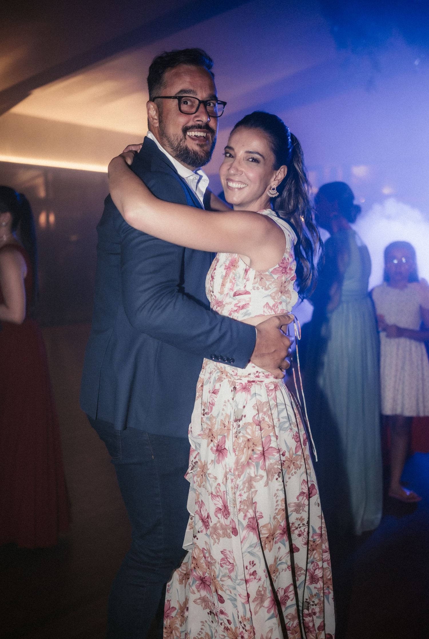 A couple dances together at an indoor event, with the woman wearing a floral dress and the man in a dark suit.