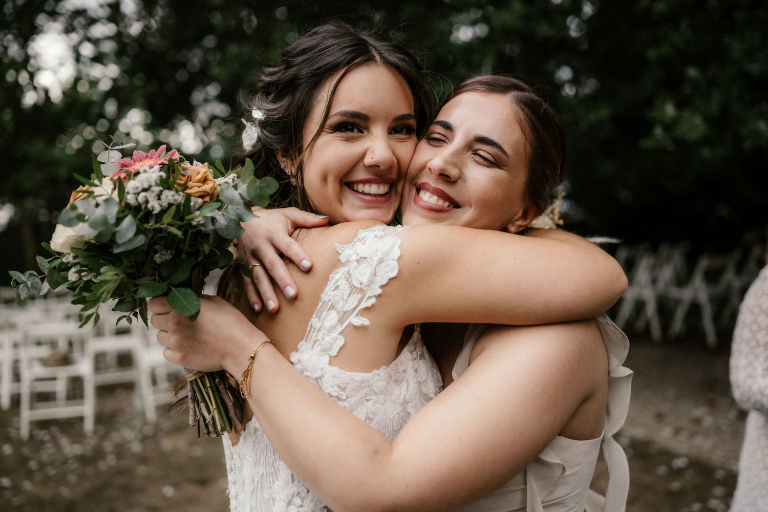 Two joyful people embrace while holding a beautiful floral bouquet at an outdoor wedding ceremony.