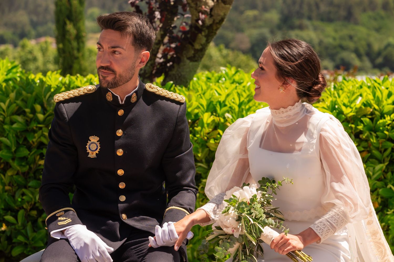 A couple in traditional wedding attire sits together outdoors with a bouquet of white flowers against lush greenery.