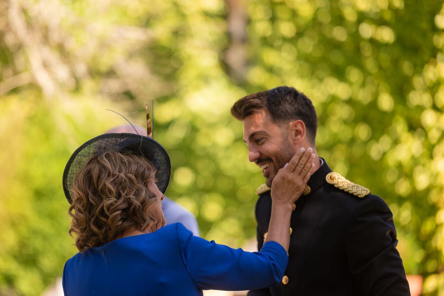 A couple embraces and kisses in a park setting with green trees and sunlight in the background.