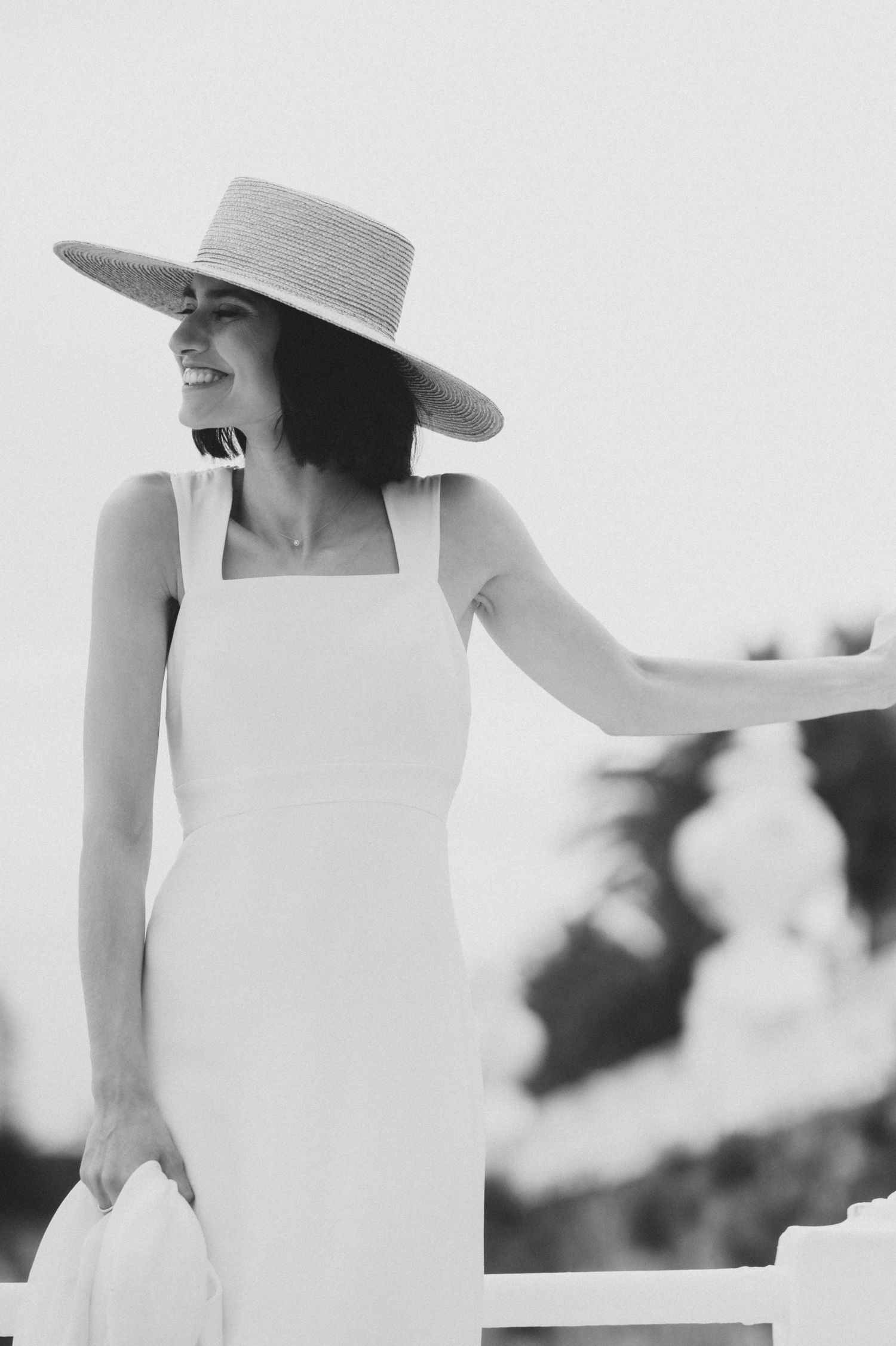 Woman in white dress and wide-brimmed hat standing elegantly in soft black and white tones.