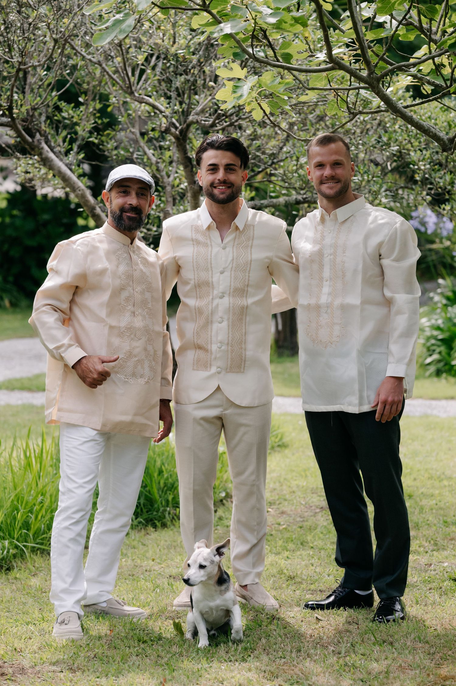 Three people in white formal attire stand together outdoors with a small white dog on grassy lawn.