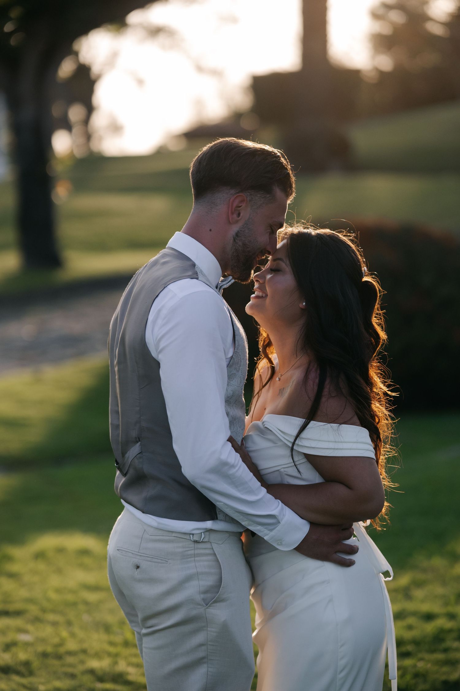 A romantic couple embraces during golden hour in a grassy outdoor setting, silhouetted by warm sunlight.