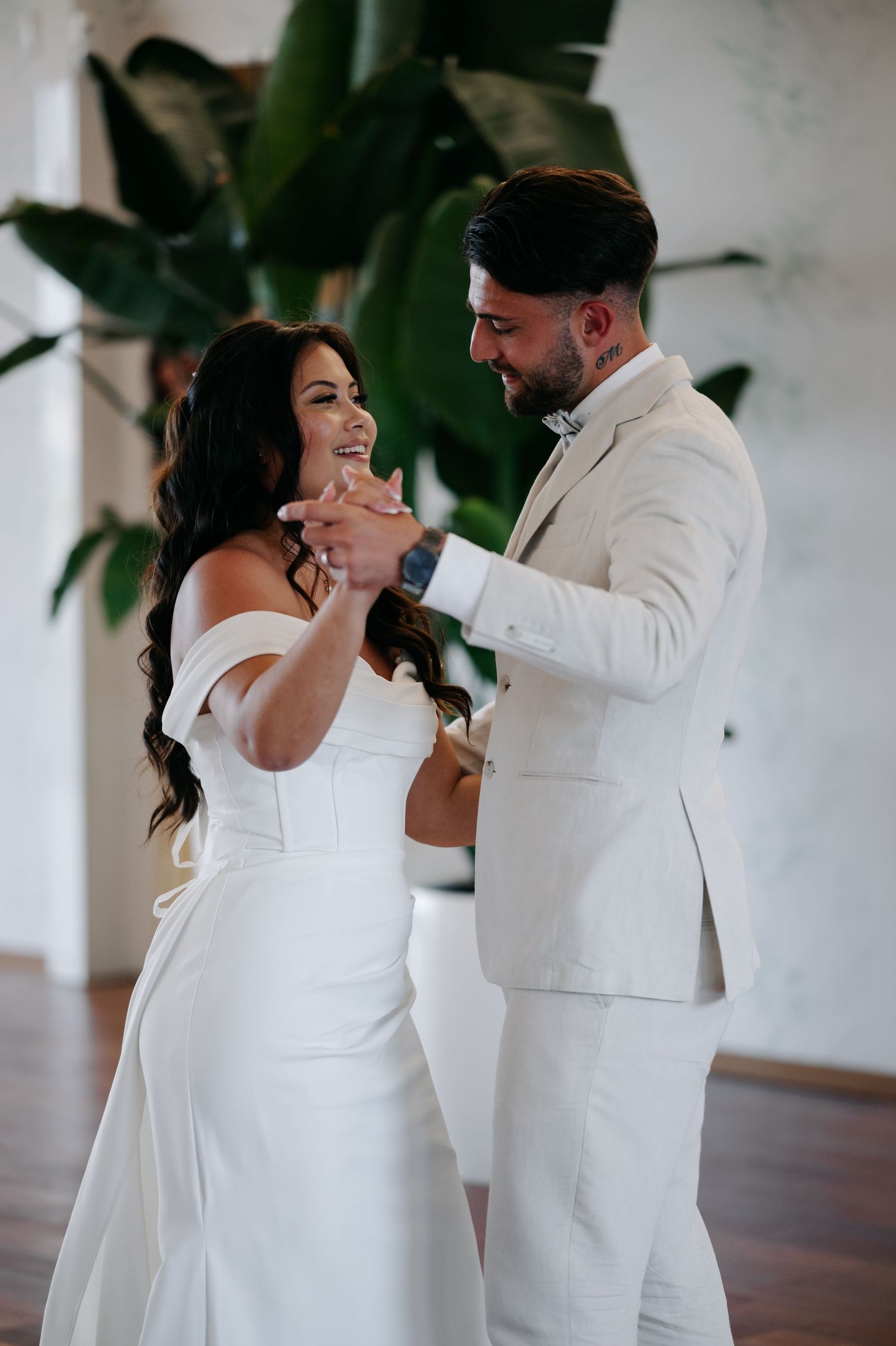A couple shares their first dance at their wedding, both dressed in elegant white attire with tropical plants in background.