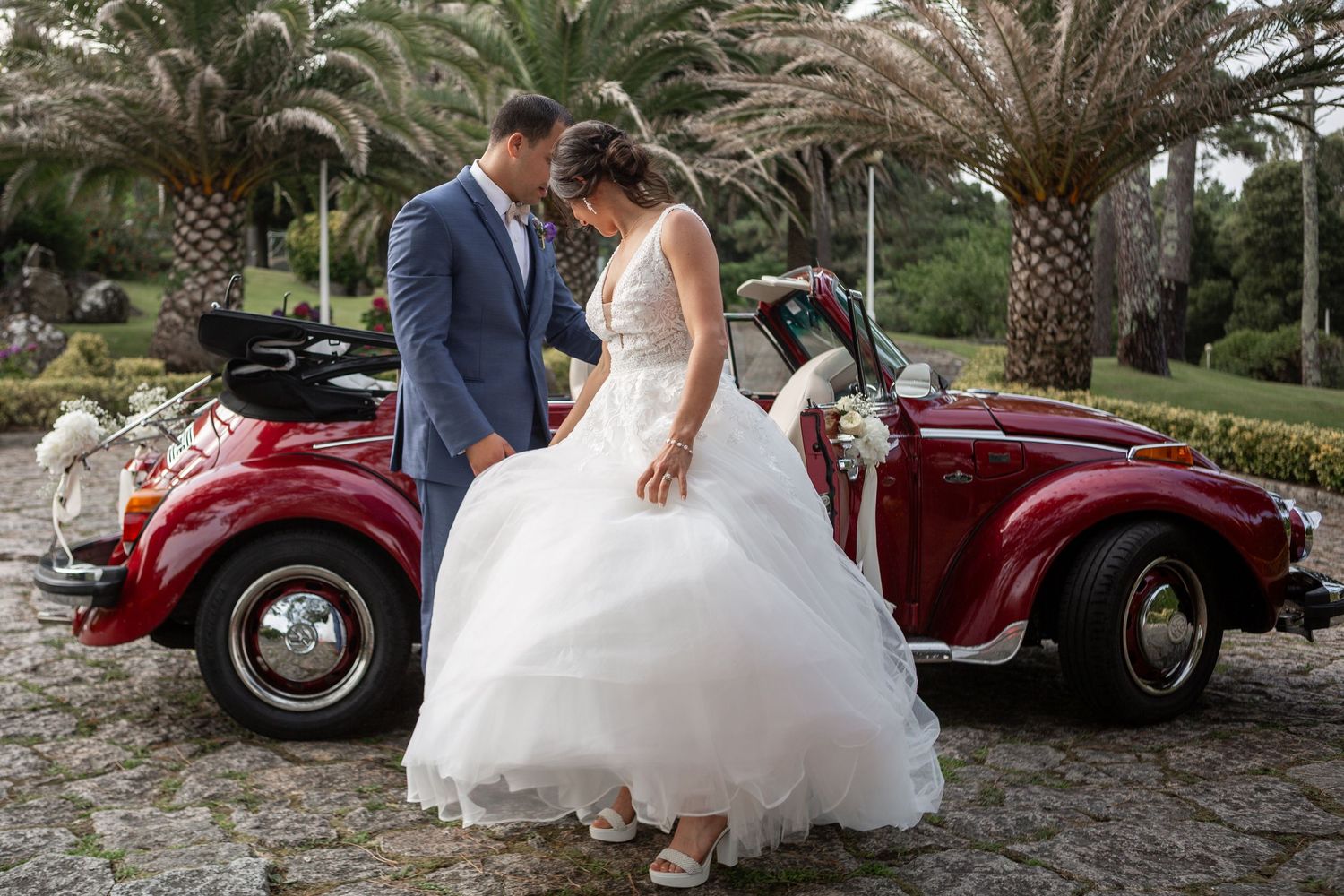 Wedding party poses with vintage red cars and motorcycles against a palm tree backdrop.