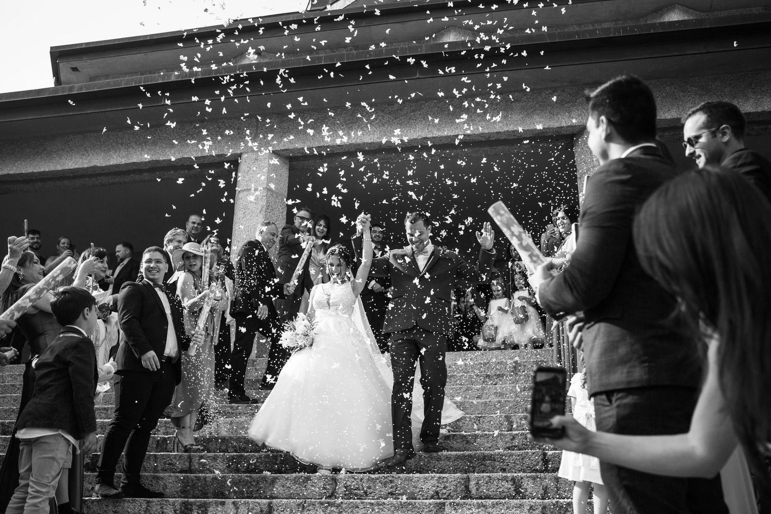 Wedding guests shower a couple with confetti as they exit the church ceremony in black and white.
