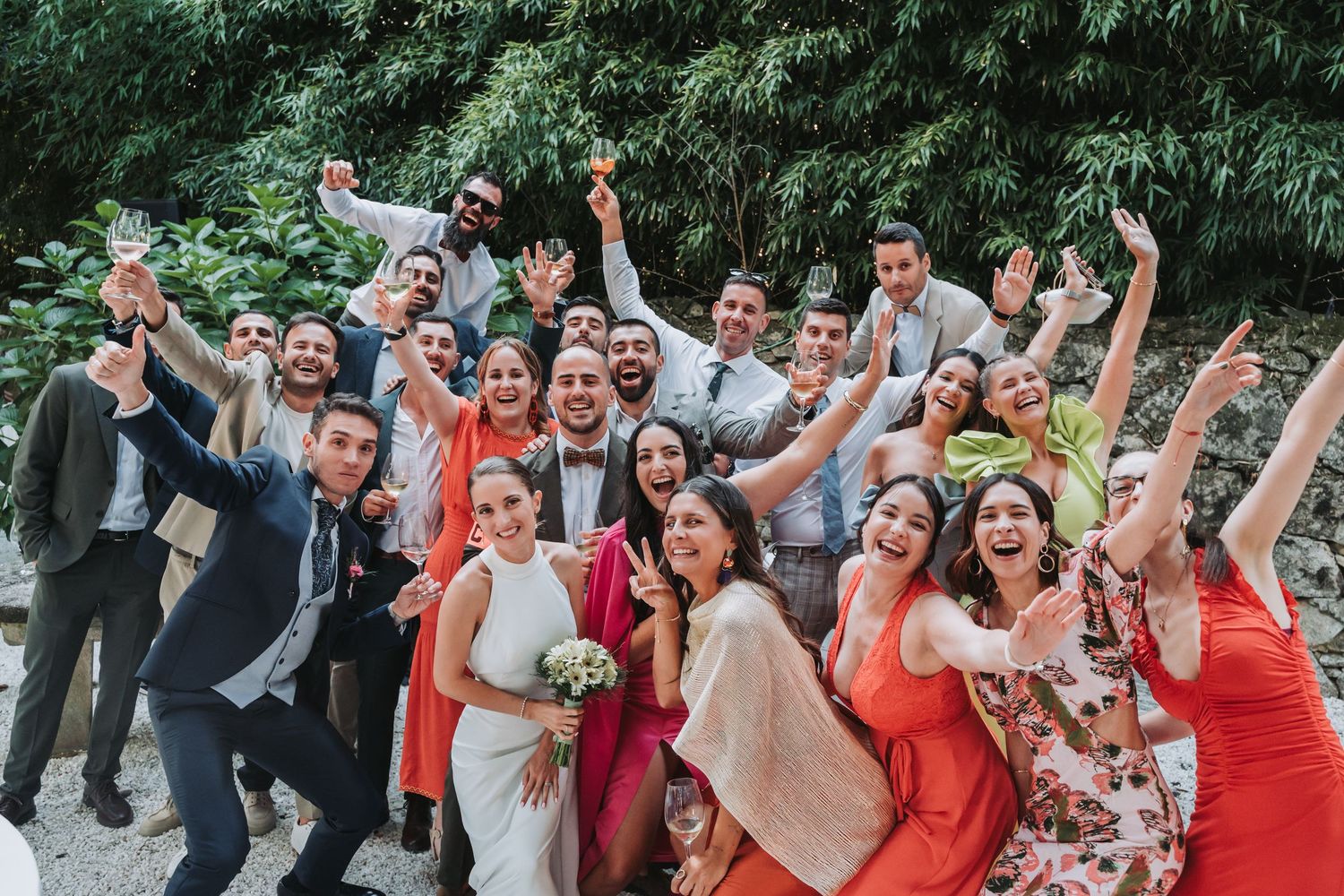 Wedding party in coral dresses and dark suits celebrate joyfully outdoors with raised arms against green foliage.