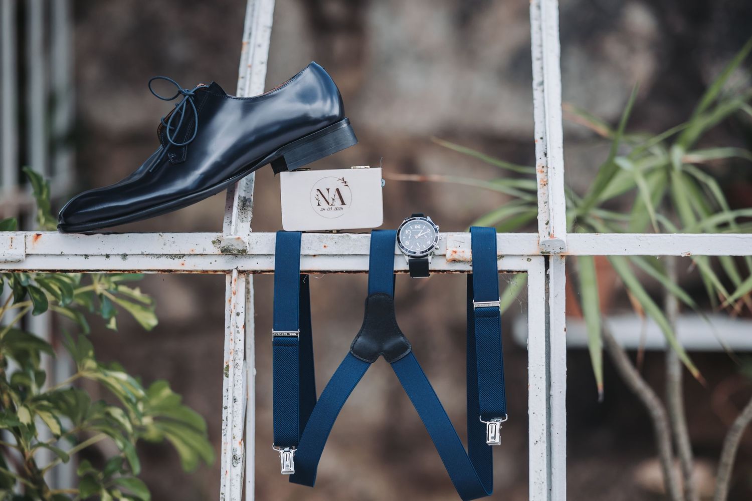 A formal black leather dress shoe and navy blue suspenders arranged on a white vintage ladder against a blurred background.
