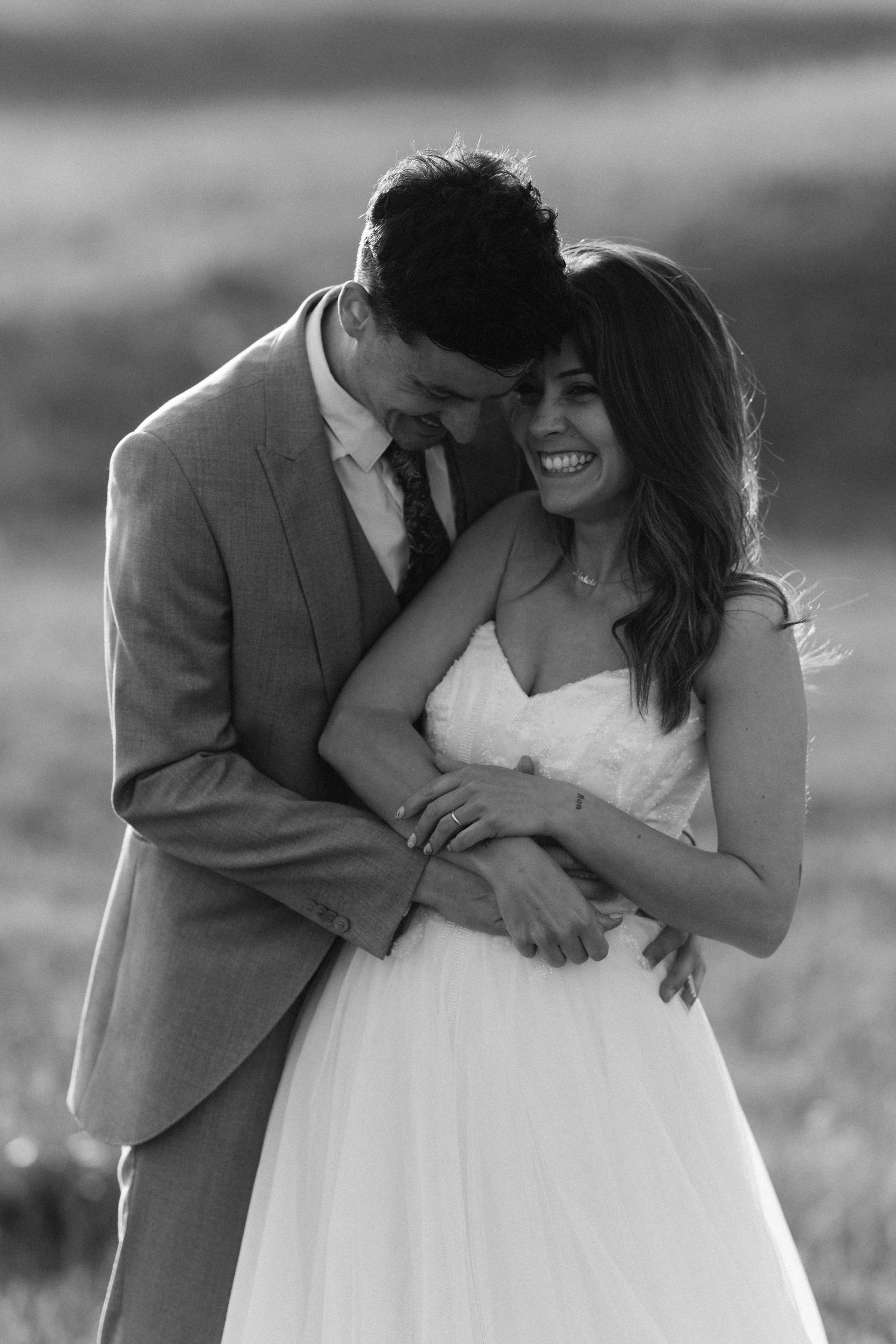 Groom embraces laughing bride from behind in joyful black and white wedding portrait outdoors.