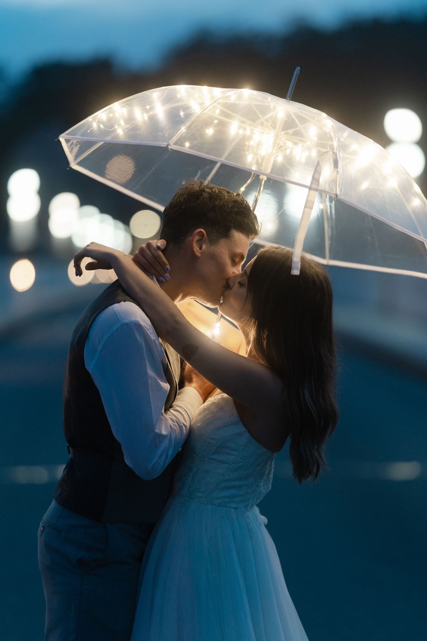 Couple kissing under a transparent umbrella with fairy lights at night, wedding photo with bokeh background.