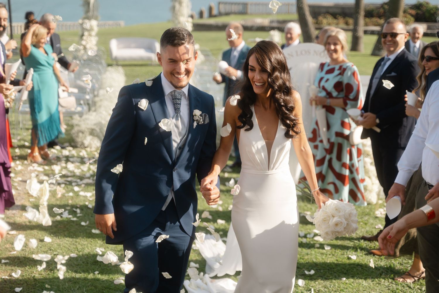 Wedding guests celebrate outdoors under white floral decorations during a sunny ceremony.